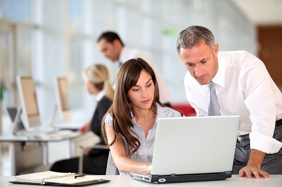 Women and men working on a laptop in a professional environment, with two blurred people using desktop computers in the background.